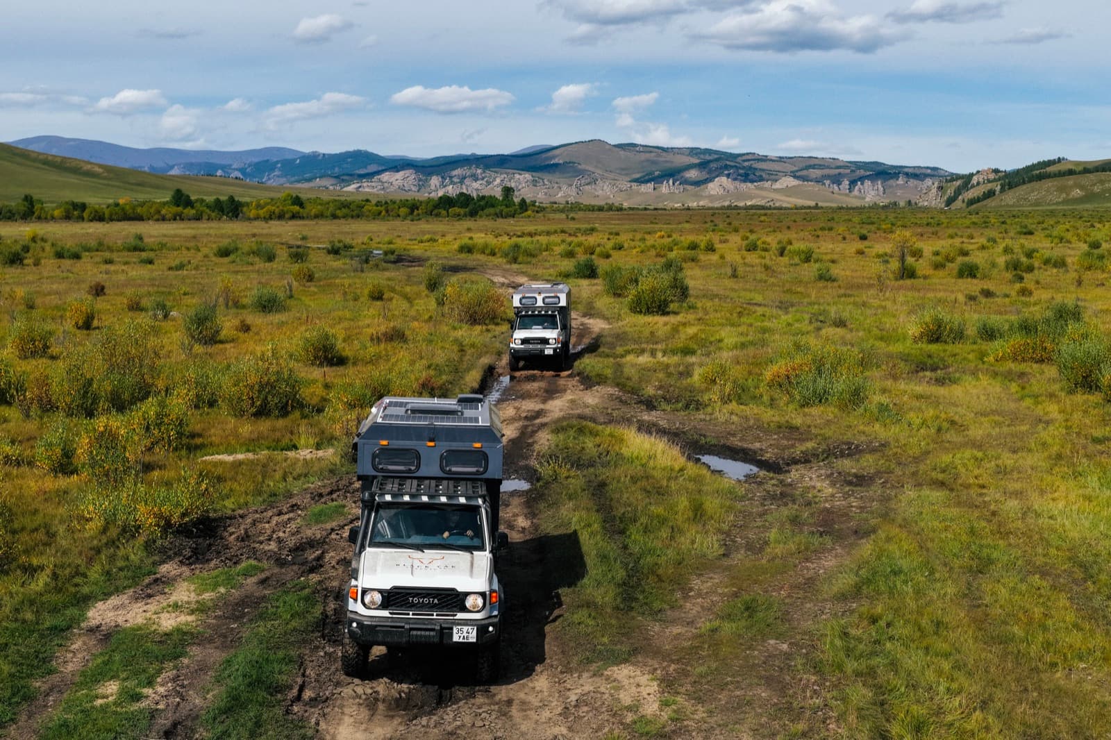 Two expedition vehicles crossing a mountain valley on a muddy trail in Mongolia